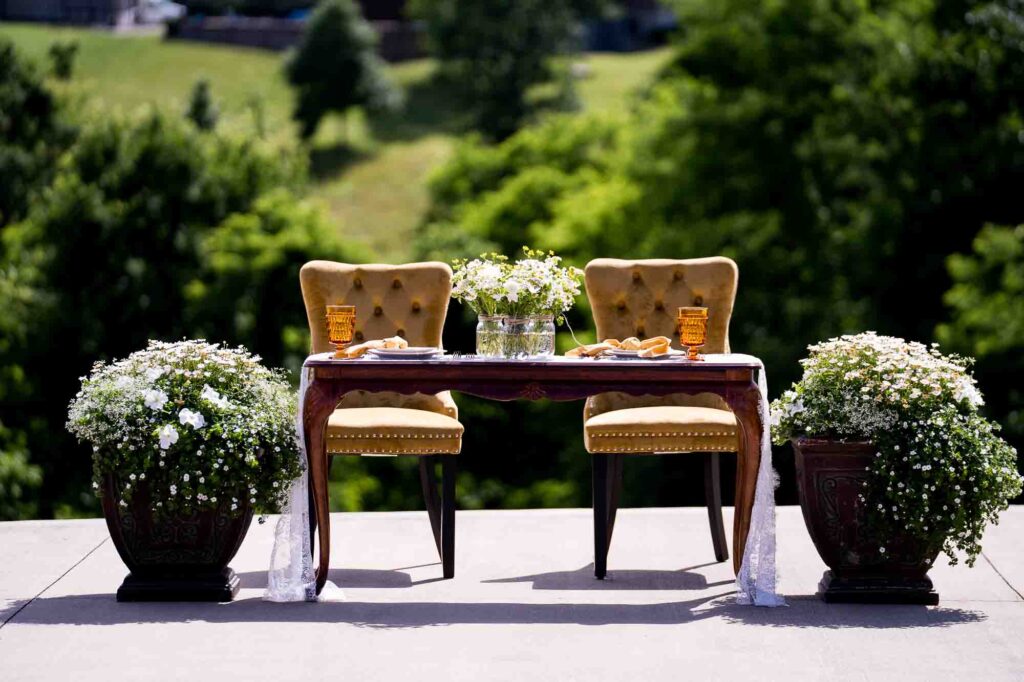 Outdoor sweetheart table for bride and groom in backyard wedding, featuring two yellow velvet chairs, large mum bushes, and a simple baby's breath centerpiece in mason jars.