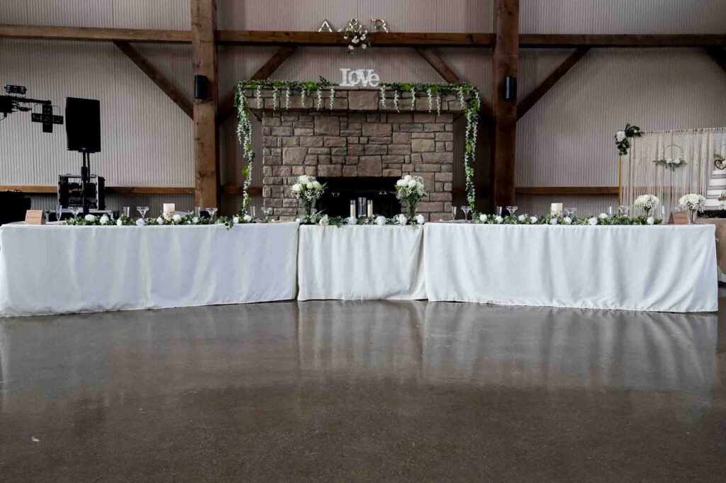 Elegant long bridal party table indoors in front of stone fireplace, with white tablecloth, white roses in glass vases, and trailing eucalyptus greenery.